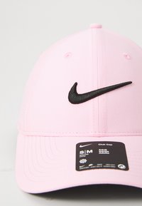 Lyserød Nike baseballcap med sort broderet swoosh-logo og et sort label på den forreste skygge, der viser størrelse og produktdetaljer.