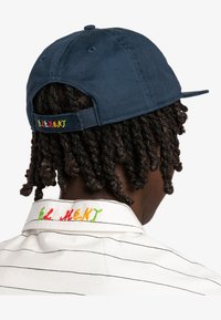Marineblauwe katoenen pet met geborduurde meerkleurige "ELEMENT" tekst op de achterkant. Gestreepte witte kraag van een shirt zichtbaar, die ook geborduurde tekst bevat.