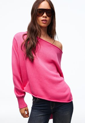 Jonge vrouw die een oversized felroze off-shoulder trui, donkere spijkerbroek en grote schildpadkleurige zonnebril draagt tegen een witte achtergrond.