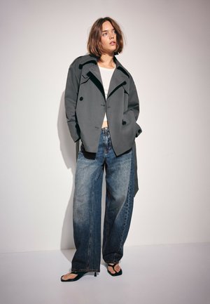 Kvinde med skulderlangt brunt hår iført en stor grå jakke, hvid crop top, jeans med vide ben og sorte sandaletter med hæl, stående mod en ensfarvet væg.