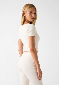 Biały krótki top z krótkimi rękawami, o gładkiej teksturze. Noszony z pasującymi jasnymi spodniami, podkreślającymi dopasowaną sylwetkę.