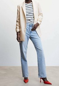 Béžový blejzr přes pruhovanou košili, světle modré rovné džíny a červené špičaté lodičky. Hladké tkaniny a ostré linie definují tento outfit.