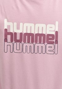 Jasnoróżowa koszulka z wyraźnym napisem "hummel" w powtarzającym się wzorze, używającym białego, różowego i ciemnoróżowego koloru na gładkiej fakturze materiału.