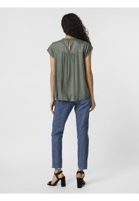 Blouse vert olive à manches courtes, détail dos en forme de goutte et corsage froncé. Associée à un jean en denim bleu et des sandales à talons noires.