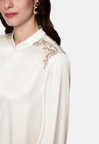 White blouse with a stand-up collar, featuring intricate light gold embroidery on the shoulder and soft fabric with a smooth texture.