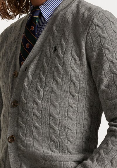 Grijze kabelgebreide cardigan van textuurstof, met houten knopen en een klein logo op de linkerborst. Gecombineerd met een gestreept shirt en stropdas.