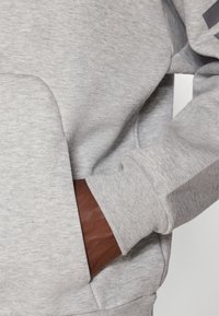 Sweatshirt cinza com uma textura suave, bolso frontal e detalhe em couro castanho na manga. Apresenta punhos canelados e um corte descontraído.