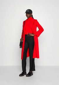 Casaco longo vermelho, gola alta vermelha justa, calças pretas ajustadas, sapatos pretos robustos e um boné preto com logótipo branco. Texturas suaves e linhas elegantes.