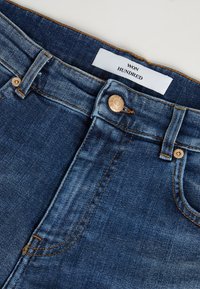 Siniset farkut korkealla vyötäröllä, joissa on vetoketjulukko ja yksi messinkipainike. Lisää etiketti "WON HUNDRED" oransseilla ommelkoristeilla.