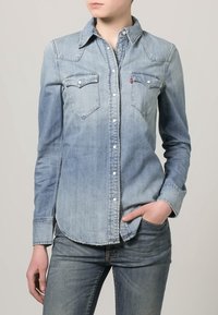 Chemise en denim bleu clair à manches longues, col pointu et deux poches poitrine. Boutons blancs et ourlet incurvé. Dégradé de couleur délavé.