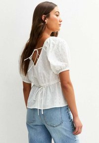 Blouse blanche en broderie anglaise avec des manches bouffantes, dotée d'un détail à nouer dans le dos. Le tissu est léger, texturé et ajusté à la taille.