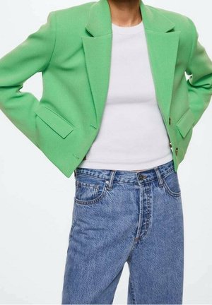 Personne portant un blazer court vert vif sur une chemise blanche, associé à un jean en denim bleu taille haute, debout les mains sur les hanches.