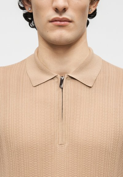 Junger Mann, der ein beiges, strukturiertes Strickshirt mit umgeschlagenem Kragen und einem kurzen silbernen Reißverschluss am Hals trägt, es sind nur Hals und unteres Gesicht sichtbar.