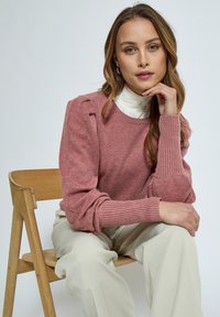Light pink knit sweater with ribbed cuffs, shoulder detailing, and a crew neckline. Worn over a white high-collared top, paired with cream trousers.