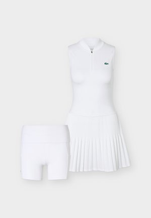 Bijela teniska haljina bez rukava sa patentnim zatvaračem i plisiranom suknjom te odgovarajućim bijelim uskim kratkim hlačicama na jednobojnoj pozadini.