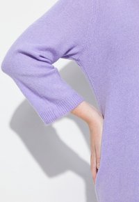 Lavendelfarbener Pullover mit dreiviertel Ärmeln, der eine weiche Textur und gerippte Bündchen aufweist. Abgebildet mit einer Hand auf der Hüfte platziert.