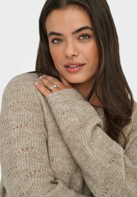 Beige adīta džemperis ar vaļīgu audumu, plašu kaklu un ribotām piedurknēm. Modelis rāda sudraba gredzenu uz rokas, ar vidēji garām brūnām matiem.