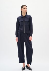 Giacca navy con zip e due tasche frontali, abbinata a pantaloni navy larghi. Il modello indossa scarpe con tacco a punte nere.
