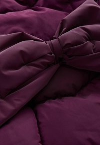 Tissu en velours bordeaux matelassé avec un détail de nœud proéminent au centre. La texture semble lisse avec des sections rembourrées.