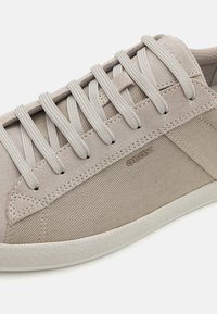 Beige sportcipő, amely velúrból és hálóból készült, lapos fűzővel, gumitalppal és varrott részletekkel. A "GEOX" logó látható az oldalon.