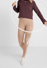 Beige Leggings mit weißen Seitenelementen und einem Logo, kombiniert mit einem dunkelmaroonen, kürzeren Sweatshirt. Das Model hält einen weißen Hula-Hoop.