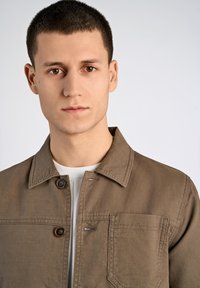 Brun bomullsjacka med spetsig krage, två framfickor och knäppning. Bärs över en vit ribbad t-shirt med rund hals.