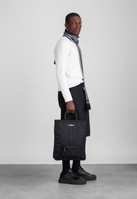 Čierna taška cez plece s prednou kapsou, obsahujúca logo. Modelka má na sebe biely sveter, šedý šál a čierne topánky. Jednoduchý dizajn, matná textura.