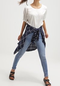 Hvid kortærmet bluse, knyttet marineblåt ternet skjorte om taljen, lyseblå skinny jeans og sorte svangsandaler.