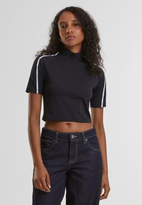 Svart kortermet t-skjorte med høy hals, med hvite side-striper. Kombinert med mørke denimjeans med synlig søm og frontlukkning med knapp.