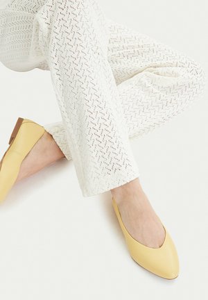 Beine übereinandergeschlagen, trägt weiße gemusterte Hosen und gelbe spitze flache Schuhe vor weißem Hintergrund.