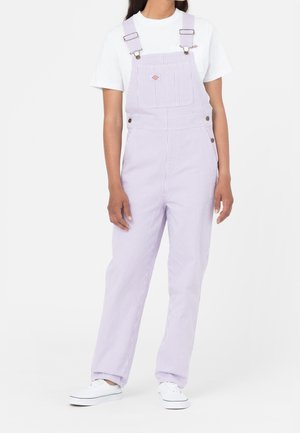 Persoon die lichtpaarse en witte gestreepte tuinbroek, wit T-shirt en witte sneakers draagt, staand tegen een effen achtergrond.