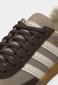 Καφέ και μπεζ αθλητικό παπούτσι Adidas με σκουρόχρωμους καφέ κορδόνια, τρεις λευκές ραμμένες ρίγες και σόλα χρώματος καραμέλας, τοποθετημένο σε γωνία για να αναδειχθούν οι λεπτομέρειες.