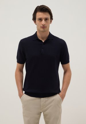 Homme portant un polo noir à manches courtes et un pantalon beige, debout les mains dans les poches devant un fond clair uni.