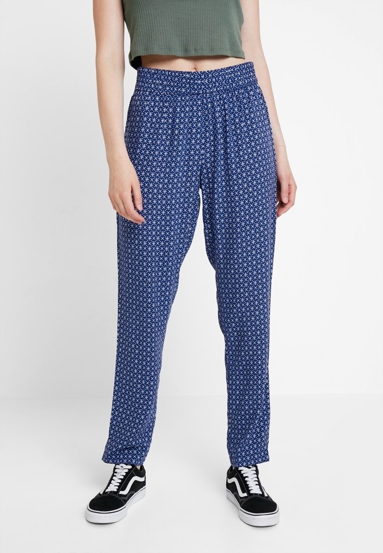 Personne portant un pantalon bleu à motifs, des baskets noires et blanches, et un top vert court sans manches sur un fond uni.