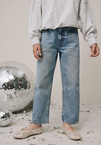 Hanorac larg gri deschis, blugi din denim albastru împodobiți cu paiete și pantofi balerini bej cu paiete, pe un fundal festiv.