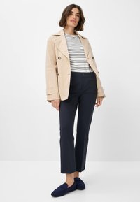 Beige doppelreihiger Trenchcoat, gestreiftes Top in Weiß und Marineblau, marineblaue maßgeschneiderte Hose und marineblaue Slipper mit flacher Sohle.
