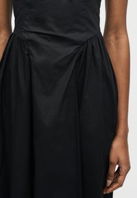 Μαύρο αμάνικο φόρεμα με συγκεντρωμένες λεπτομέρειες υφάσματος στη μέση, που δείχνει μέρος του δεξιού χεριού και του βραχίονα του μοντέλου, σε ένα απλό φόντο.