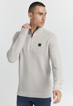 Suéter gris claro con un cuello de media cremallera, textura acanalada y un pequeño parche de logo negro en el pecho; combinado con unos jeans azul oscuro.