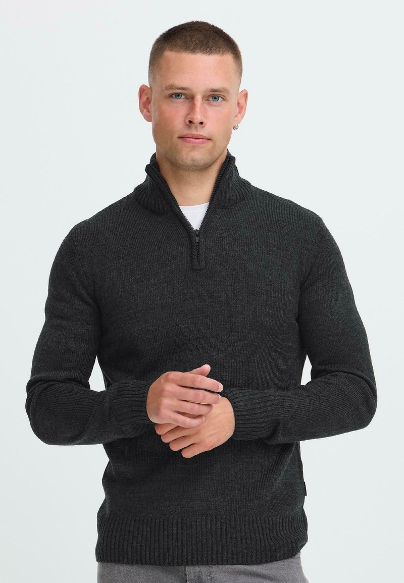 Mann med kort hår iført en mørk grå half-zip genser og lysegrå bukser, som står vendt mot kameraet med hendene foldet foran seg.