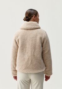 Beżowa kurtka z futra syntetycznego o miękkiej, teksturowanej powierzchni, z szerokim kołnierzem i długimi rękawami, charakteryzująca się luźnym krojem i zaokrąglonym dołem.