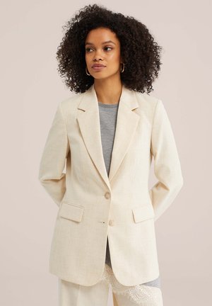 Göndör hajú nő krémszínű blézerben, szürke csipkés aljú felsőt viselve, háttal a kezével a hát mögött egy bézs háttér előtt áll.