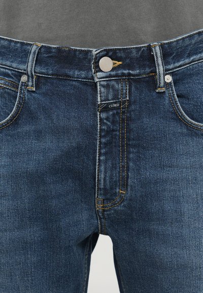 Jeans din denim albastru închis, cu un croi slim, având cusături galbene, fermoar la brâu și închidere cu un buton metalic. Eticheta brandului este vizibilă pe față.