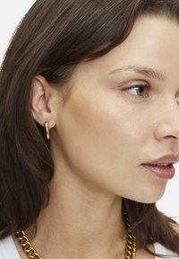 Orecchino a cerchio dorato esposto all'orecchio di un modello, caratterizzato da un design liscio e curvo. I capelli sono castani scuri e il tono della pelle appare chiaro.