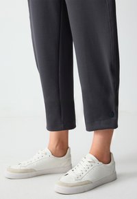 Graue cropped Hose mit seitlichen Nähten und schwarzen Akzentknöpfen, kombiniert mit weißen Turnschuhen, die graue Wildlederelemente und Perforationen aufweisen.