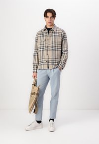 Beige plaid flannel shirt, black t-shirt, light blue trousers, white sneakers, carrying a tan tote bag with strap and front pocket.