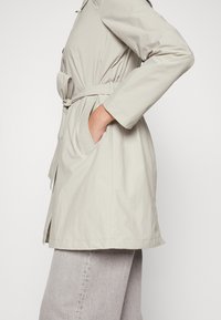 Trench-coat beige clair avec une taille ceinturée, des poches latérales et des manches longues, fabriqué dans un tissu lisse légèrement texturé.