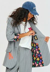 Šedý blazer s pruhy a barevně květinově podšité rukávy a vnitřek. Bílá košile s vyšitým květem a grafickým prvkem oranžového humra. Námořnická čepice.