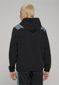 Fekete kapucnis dzseki világoskék vállrészletekkel. Rugalmas mandzsettákkal, laza szabással és sima textúrával rendelkezik.