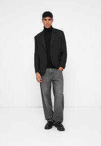 Blazer preto, suéter de gola alta preto, jeans largos cinzas e sapatos pretos usados por um modelo, contra um fundo branco liso.
