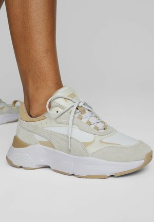 Noha v béžové a bílé tenisce Puma s texturovanou podrážkou, šněrování uvázané, na jednobarevném světlém šedém pozadí.